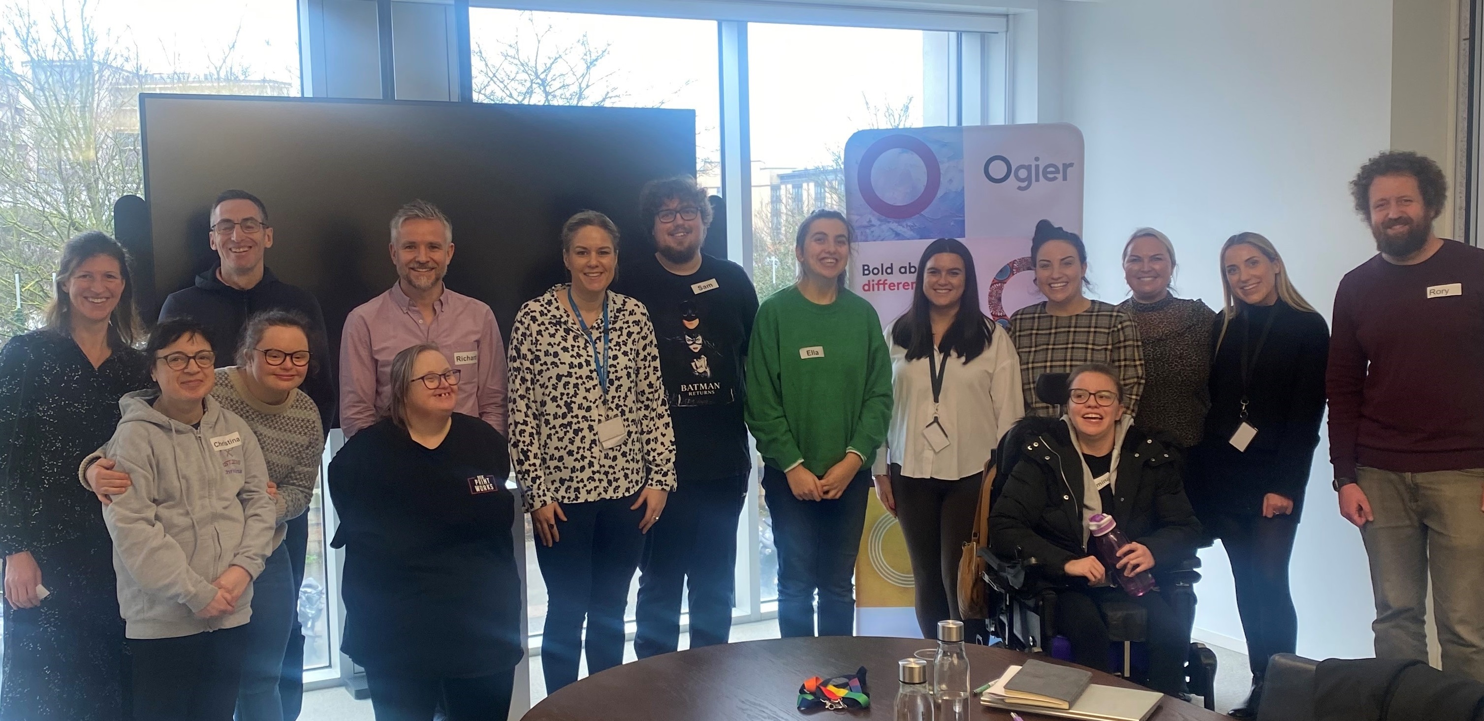 Ogier employees and six Beresford Street Kitchen crew members stand together in the Jersey office with BSK and Ogier banners behind them, celebrating partnership and workplace training initiative